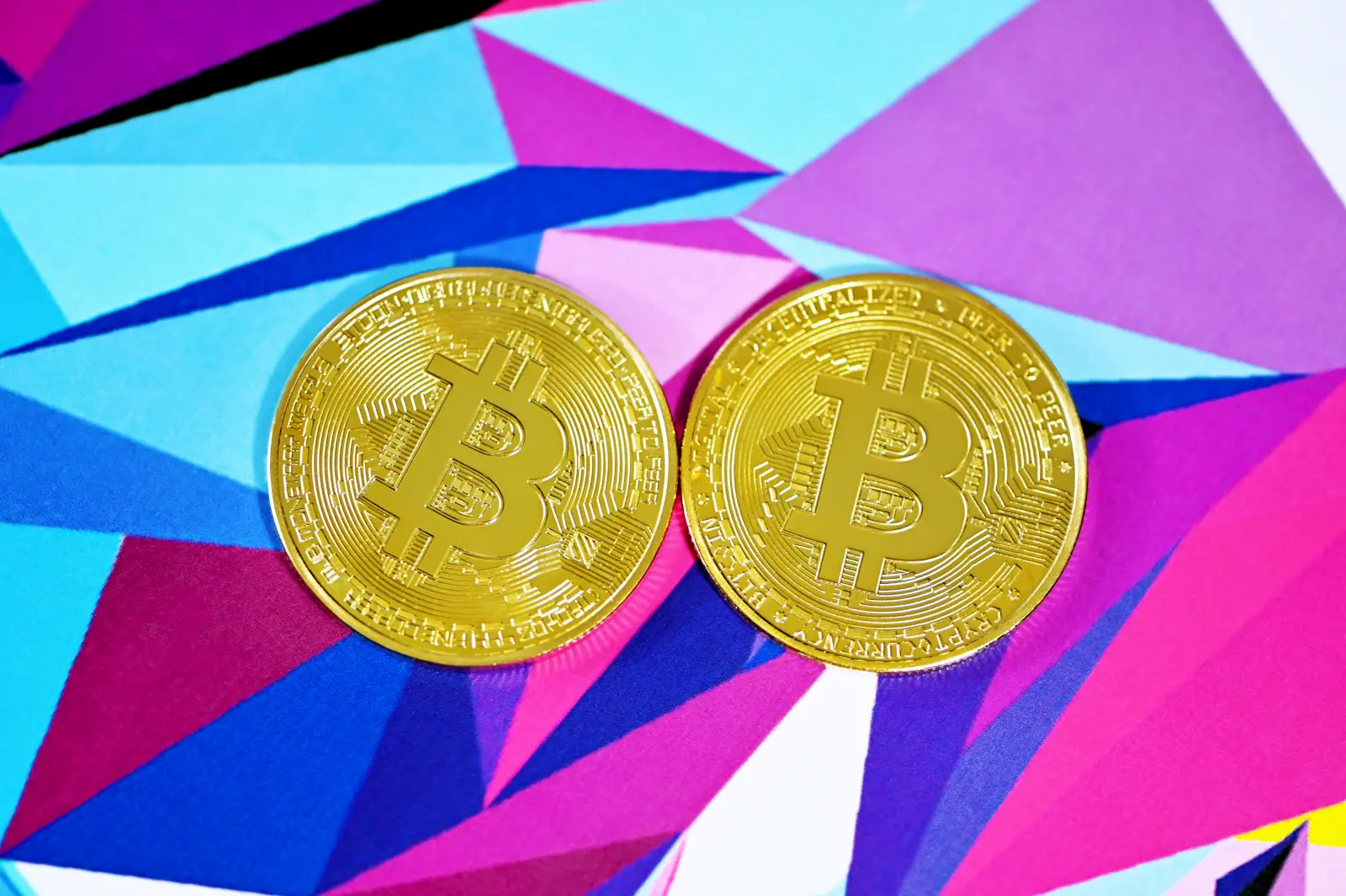 Two gold Bitcoin tokens on a vivid magenta, turquoise, and blue geometric background