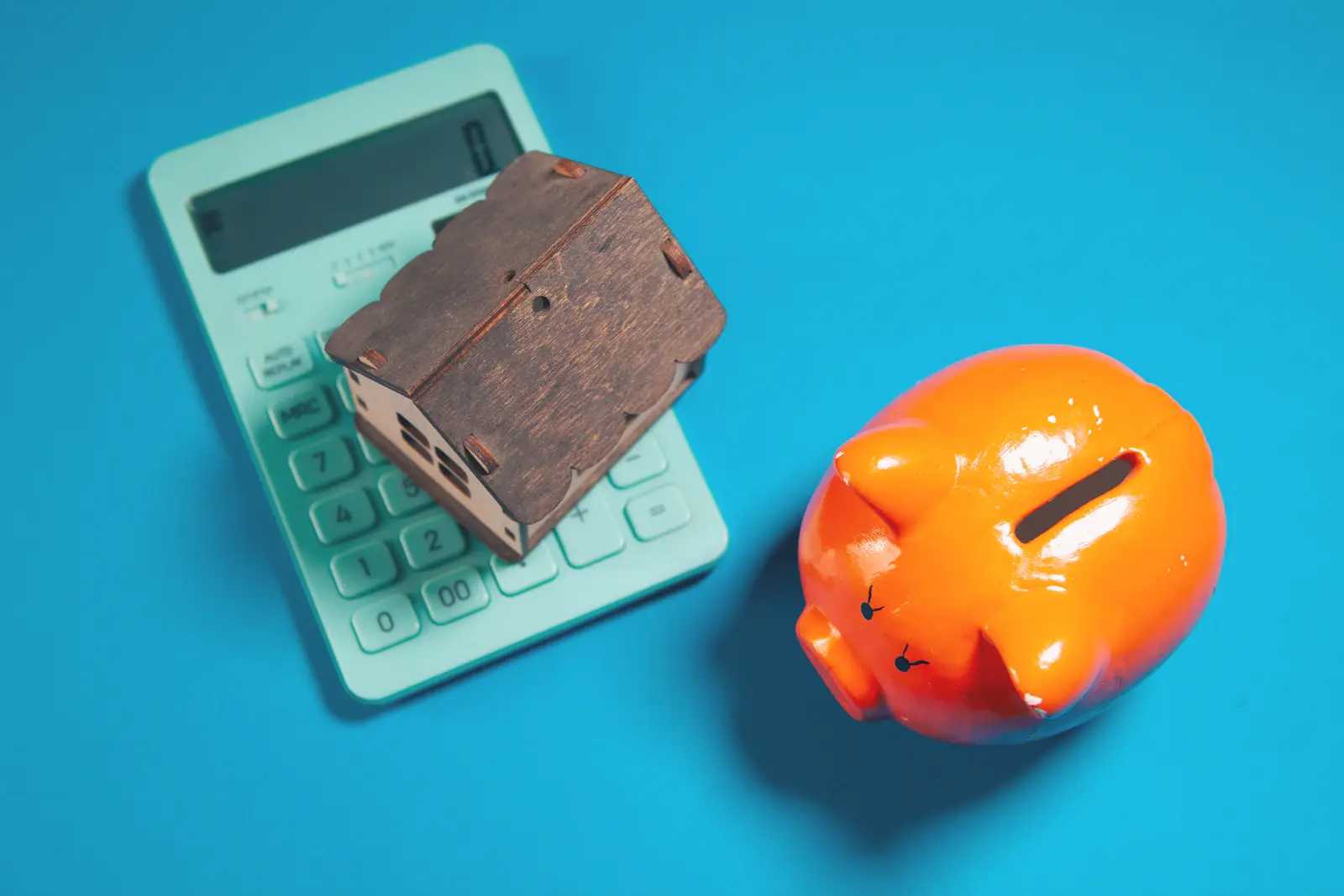 A calculator and wooden house model beside an orange piggy bank on a bright blue background